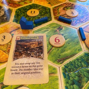 May include: A board game setup featuring a hexagonal tile board with various terrain illustrations and numbered circular tokens. A rectangular card titled Earthquake is placed on the board, featuring text and an illustration. Blue wooden game pieces are scattered.