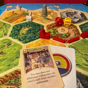 May include: The Settlers of Catan board game featuring a hexagonal tile map, red and white wooden player pieces, a grey wooden figure, two dice, and a Neighbours card with the text: You may place one settlement within only one road length from another settlement.