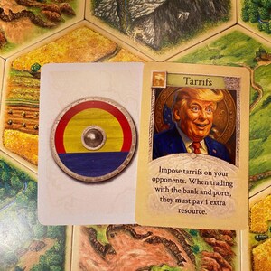 K&ouml;nnte beinhalten: Eine rechteckige Spielkarte mit einem runden Schild mit roten, gelben und blauen horizontalen Streifen und einem silbernen Buckel in der Mitte, neben einer Karte mit der Aufschrift Tarrifs, die einen Mann im Anzug zeigt.