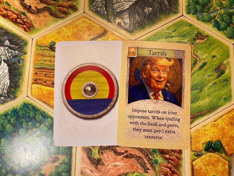 K&ouml;nnte beinhalten: Eine rechteckige Spielkarte mit einem runden Schild mit roten, gelben und blauen horizontalen Streifen und einem silbernen Buckel in der Mitte, neben einer Karte mit der Aufschrift Tarrifs, die einen Mann im Anzug zeigt.