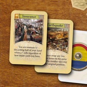May include: Board game cards with illustrations and text, including "Insurance" and "Earthquake" cards. The "Insurance" card depicts a market scene, while the "Earthquake" card shows a battle scene. Other game components are also visible.