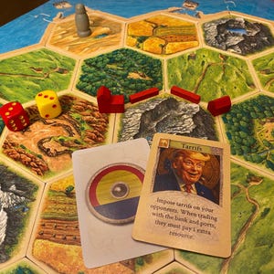 K&ouml;nnte beinhalten: Ein Brettspiel mit hexagonalen Gel&auml;ndefeldern, roten Holzh&auml;usern und Stra&szlig;en, einer grauen Spielfigur und zwei W&uuml;rfeln. Zwei Karten liegen auf dem Spielbrett, eine zeigt einen runden Schild und die andere ein Portr&auml;t mit dem Text Tarrifs.