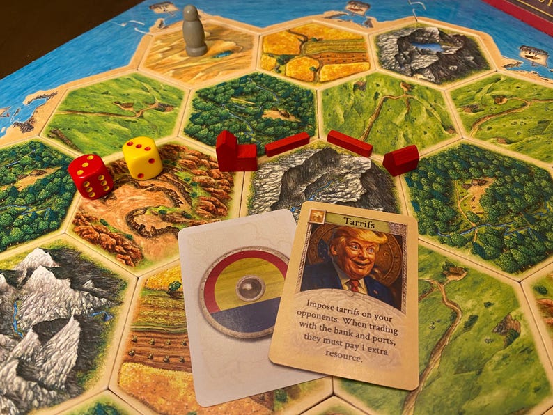 K&ouml;nnte beinhalten: Ein Brettspiel mit hexagonalen Gel&auml;ndefeldern, roten Holzh&auml;usern und Stra&szlig;en, einer grauen Spielfigur und zwei W&uuml;rfeln. Zwei Karten liegen auf dem Spielbrett, eine zeigt einen runden Schild und die andere ein Portr&auml;t mit dem Text Tarrifs.