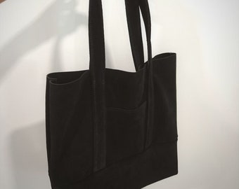 May include: A black suede tote bag with a single pocket on the front. The bag has two straps and is held by a hand.