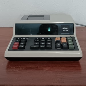 Op de afbeelding: Een vintage Royal 242PD rekenmachine in beige en zwart. Het display toont het getal 0. De toetsen zijn zwart, met rode, oranje en groene accenten. De rekenmachine staat op een houten oppervlak.