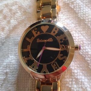 May include: Gold-tone wristwatch with a black face and gold-tone Roman numeral hour markers. The watch has a heart-shaped design in the centre of the face and the word "Betsyville" written below the heart.