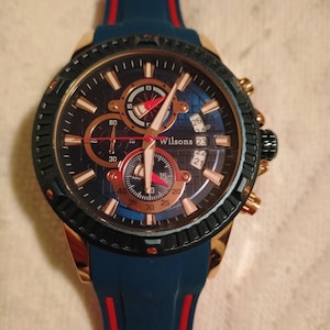 May include: A blue and red silicone wristband watch with a gold-tone case and black face. The watch has a chronograph function and a date window. The watch face has the word "Wilsons" printed on it.