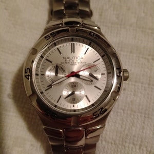 May include: A silver Nautica men's watch with a white face and black hands. The watch has a date window and a chronograph. The watch is water resistant to 100 metres.