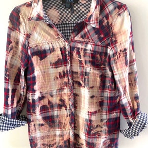 May include: A red, white and blue plaid flannel shirt with a bleach-splattered design. The shirt has a button-up front and long sleeves. The brand label "FDJ" is visible on the collar.