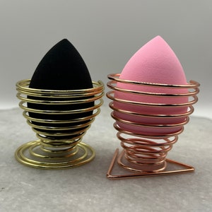 May include: Two makeup sponges, one black and one pink, are displayed in gold and rose gold metal holders. The holders are shaped like a spiral and have a triangular base.