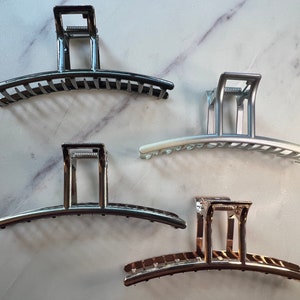 Puede incluir: Cuatro pinzas para el pelo en diferentes colores: negro, plateado, blanco y dorado. Las pinzas son de metal y tienen una forma curva con una fila de dientes.