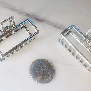 Puede incluir: Dos pinzas para el pelo de metal plateado, cada una con forma rectangular y un mecanismo de resorte. Las pinzas son aproximadamente del tamaño de una moneda de 25 centavos de dólar estadounidense.