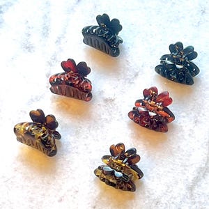 May include: Six small, flower-shaped hair claw clips in shades of brown, amber, and black are arranged on a white, textured surface. The clips have a tortoiseshell pattern and are made of a translucent material. The claw clips are a hair accessory.