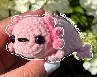 May include: A handmade pink crochet axolotl with black eyes and pink gills. The plush toy is held in a hand against a background of white flowers and green foliage. The text "LEMONYCKROCHET" is visible.