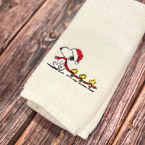 May include: White hand towel with an embroidered design of Snoopy wearing a Santa hat and a red scarf, walking with three yellow birds.