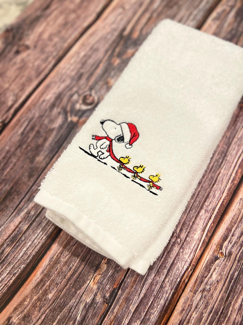 May include: White hand towel with an embroidered design of Snoopy wearing a Santa hat and a red scarf, walking with three yellow birds.