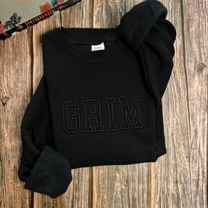 May include: Black sweatshirt with the word "GRIM" embroidered in black on the front.