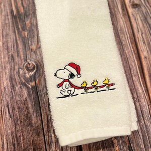 May include: White kitchen towel with an embroidered design of Snoopy wearing a Santa hat and a red scarf, pulling a sled with three yellow birds.