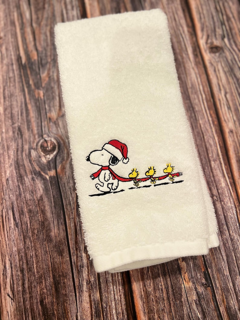 May include: White kitchen towel with an embroidered design of Snoopy wearing a Santa hat and a red scarf, pulling a sled with three yellow birds.