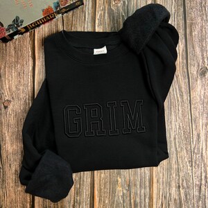 May include: Black sweatshirt with the word "GRIM" embroidered in black on the front.