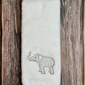 Puede incluir: Toalla de mano blanca con un elefante gris bordado con pies naranjas. La toalla está sobre un fondo de madera.