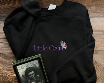May include: Black crewneck sweatshirt with the embroidered text "Little Osha" and a butterfly. A Kindle e-reader displaying the book cover of "Quicksilver" by Callie Hart rests beside the sweatshirt.