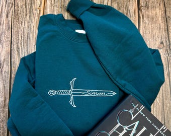 Puede incluir: Sudadera de color turquesa con un diseño de espada bordada en blanco y el nombre "Simon". Un libro con el título "Callie Hart" está al lado de la sudadera. Los artículos están sobre una superficie de madera.