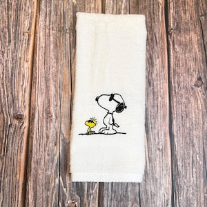 May include: White hand towel with an embroidered image of Snoopy in sunglasses and Woodstock. The black and yellow characters are on a white, rectangular towel. The towel is made of a soft, absorbent material.