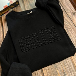 May include: Black sweatshirt with the word "GRIM" embroidered in a large, bold font.