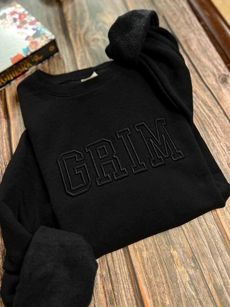May include: Black sweatshirt with the word "GRIM" embroidered in a large, bold font.