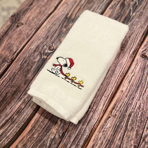 May include: White kitchen towel with an embroidered image of Snoopy wearing a red Santa hat and pulling a sled with four yellow birds.