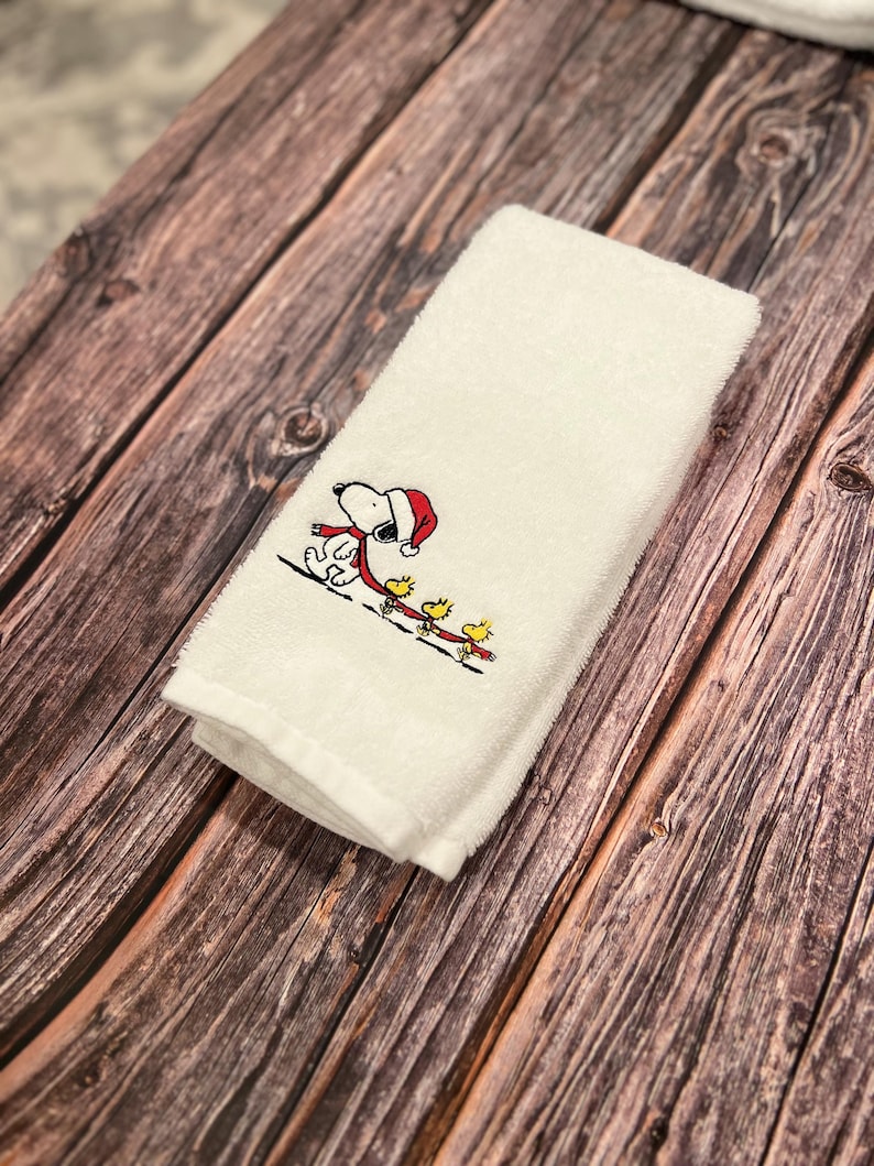 May include: White kitchen towel with an embroidered image of Snoopy wearing a red Santa hat and pulling a sled with four yellow birds.
