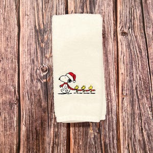May include: White kitchen towel with an embroidered design of Snoopy wearing a Santa hat and pulling a string of yellow birds.