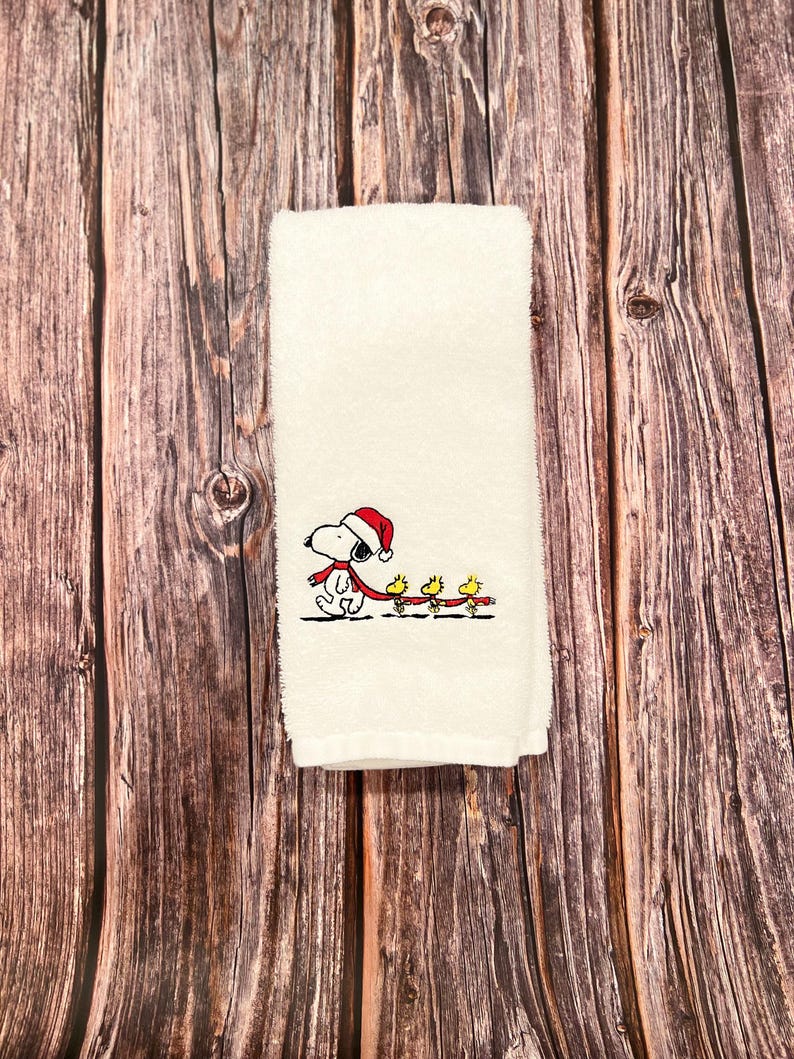 May include: White kitchen towel with an embroidered design of Snoopy wearing a Santa hat and pulling a string of yellow birds.
