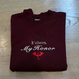 Puede incluir: Sudadera color burdeos con texto bordado en blanco que dice "It's been. My Honor." y un gr&aacute;fico de drag&oacute;n rojo.