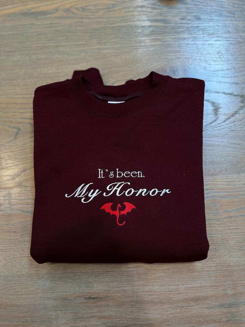 Puede incluir: Sudadera color burdeos con texto bordado en blanco que dice "It's been. My Honor." y un gr&aacute;fico de drag&oacute;n rojo.