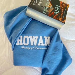 Op de afbeelding: Een blauwe hoodie met de woorden "ROWAN Daddy of Terrasen" geborduurd op de voorkant. Een paperback boek getiteld "Kingdom of Ash" van Sarah J. Maas ligt op de hoodie.