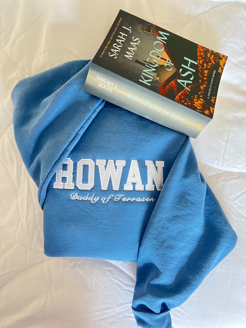 Op de afbeelding: Een blauwe hoodie met de woorden "ROWAN Daddy of Terrasen" geborduurd op de voorkant. Een paperback boek getiteld "Kingdom of Ash" van Sarah J. Maas ligt op de hoodie.