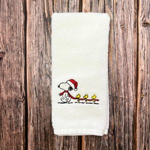 May include: White hand towel with an embroidered design of Snoopy wearing a Santa hat and pulling a sled with three yellow birds.