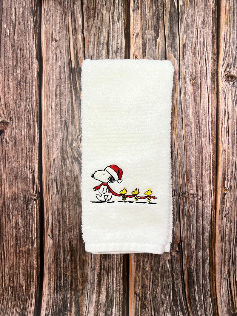 May include: White hand towel with an embroidered design of Snoopy wearing a Santa hat and pulling a sled with three yellow birds.