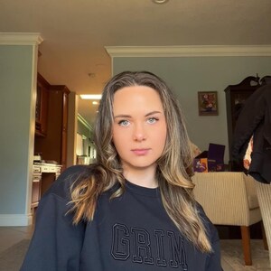May include: A woman with long blonde hair is wearing a black sweatshirt with the word "GRIM" embroidered on the front in white.