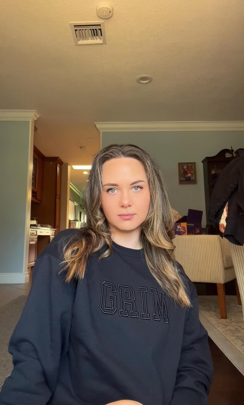 May include: A woman with long blonde hair is wearing a black sweatshirt with the word "GRIM" embroidered on the front in white.