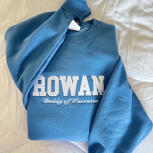Op de afbeelding: Een blauw sweatshirt met witte geborduurde tekst die "ROWAN Daddy of Terrassen" luidt.