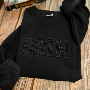 May include: Black sweatshirt with the word "GRIM" embroidered in black on the front.