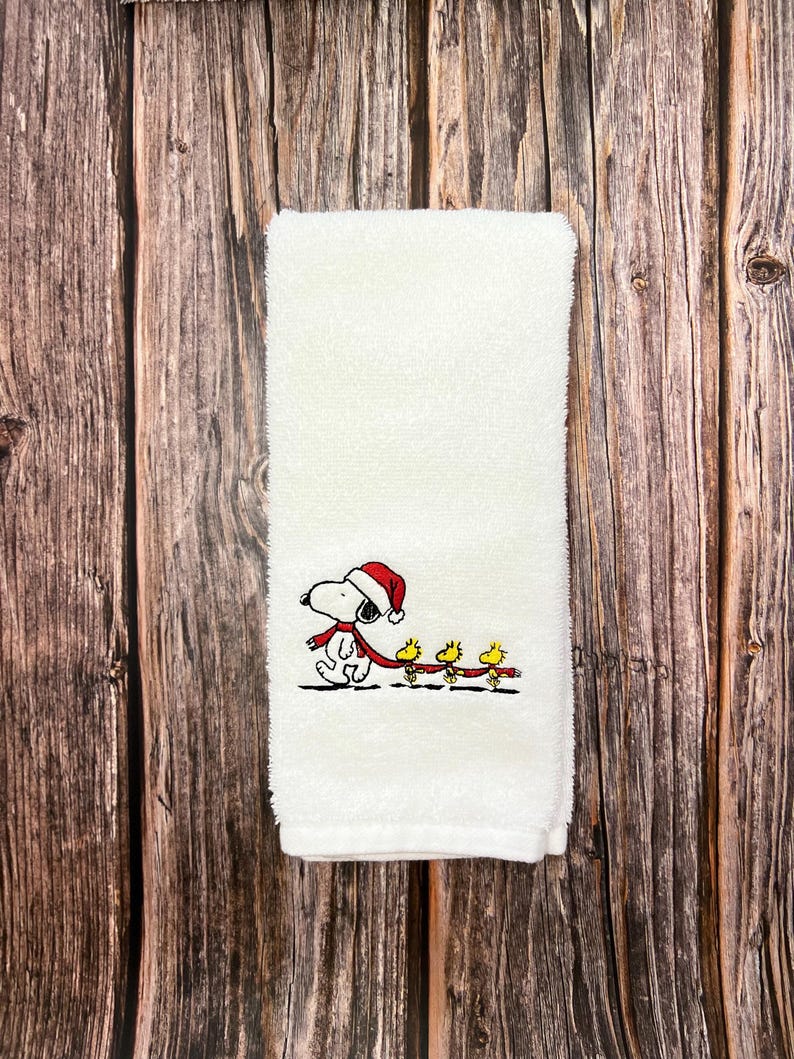 May include: White kitchen towel with an embroidered design of Snoopy wearing a Santa hat and pulling a string of yellow birds with red scarves.