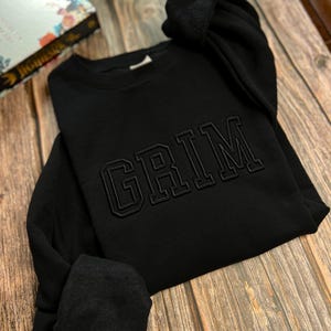 May include: Black sweatshirt with the word "GRIM" embroidered in a large, bold font.