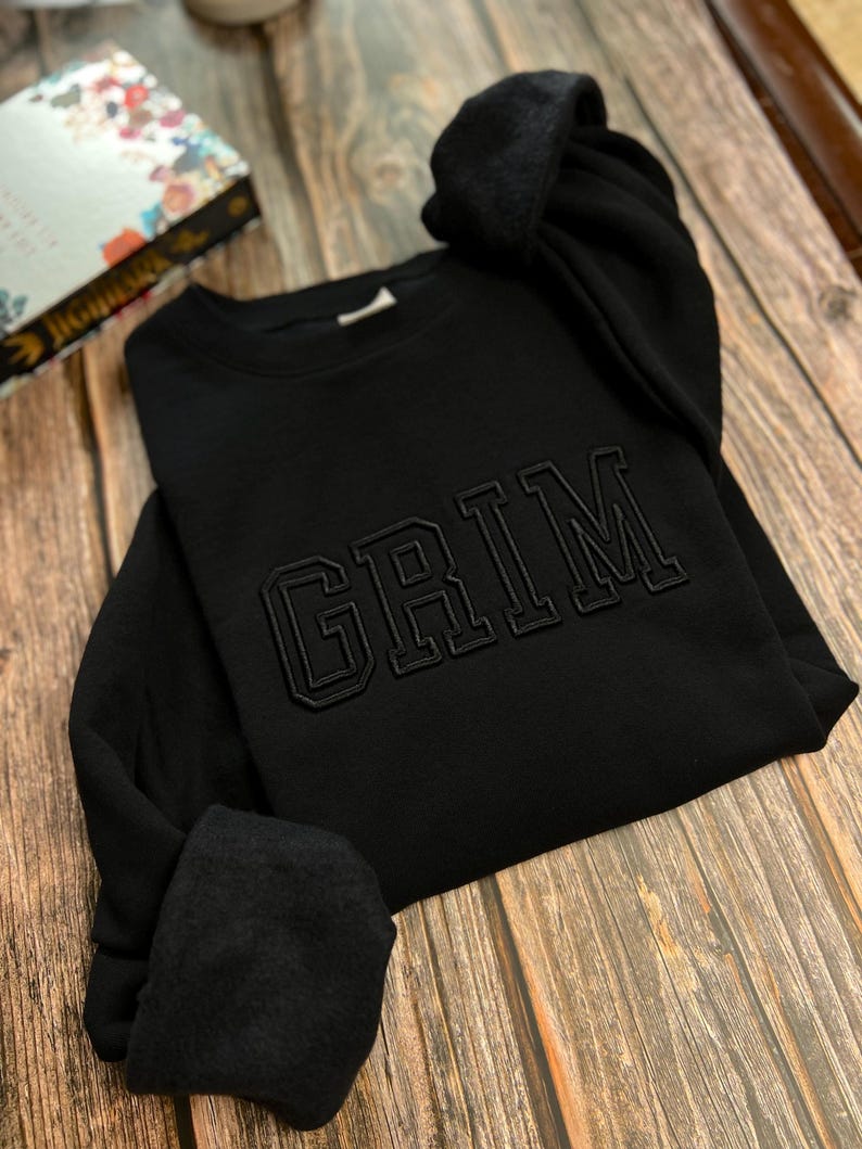 May include: Black sweatshirt with the word "GRIM" embroidered in a large, bold font.