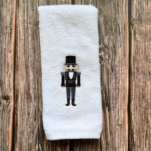 May include: White hand towel with an embroidered nutcracker design. The nutcracker is wearing a black hat, black jacket, and black pants. The nutcracker has a white face with a black mustache.