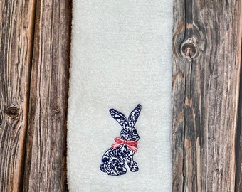 以下が含まれることがあります： ピンクのリボンをつけた青と白の刺繍のウサギが描かれた白いハンドタオルです。
