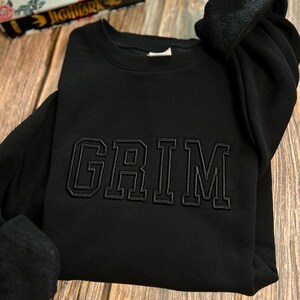 May include: Black sweatshirt with a black embroidered word "GRIM" on the front.